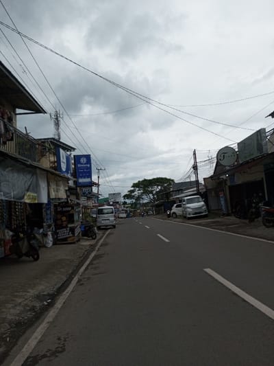 The image presents a street scene in the city of Melakudikadu, India, viewed from a low angle. The sky is overcast with a dense layer of grey clouds. A two-lane asphalt road, marked with white solid and dashed lines, occupies the center and foreground, extending into the distance with a slight upward incline.
On the left side of the road, a sidewalk fronts a continuous line of multi-story buildings, consistent with commercial establishments or residences with ground-floor shops. Some shops feature awnings or tarpaulin coverings. Specific visible elements include a sign displaying "Rp 15.000" and another small stall sign indicating "Rp 3000" and "RIGOL'S EXTRA PEDAS ORIGINAL." Two prominent blue vertical signs are affixed to poles; one displays the "Bintang" logo with Arabic script underneath, and another below it bears the text "UPLINK." A tall cellular communication tower is visible behind the buildings on this side. Several motorcycles, including a scooter, are parked along the sidewalk. A silver utility minivan is also parked on the shoulder or sidewalk.
On the right side of the road, buildings continue the commercial frontage. A circular white sign with the text "TIARA" in a stylized font, accompanied by "Tiara Wedding" below it, is visible on one building. Parked vehicles on this side include a white compact hatchback and a silver minivan. Further down the street, additional motorcycles are parked.
The road itself appears clear of active traffic in the immediate and mid-ground. Power lines are visible crisscrossing the upper portion of the frame. Green foliage from trees is visible in the background beyond the urban structures. No individuals are clearly discernible performing specific actions or interactions within the scene, although indistinct figures may be present in the distant background.