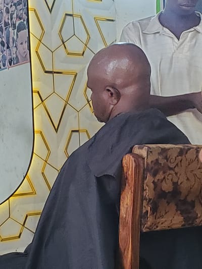 This media file shows two people in what appears to be a barber shop in Ampan, Nigeria. One individual, likely a barber, is wearing a light-colored shirt with vertical stripes. The other individual, a customer, is bald and wearing a black garment. The customer's head is the primary focus of the image, tilted to the side.

The background features a distinctive geometric tile pattern with gold accents, suggesting a modern or decorative interior. A portion of a patterned upholstered object, possibly a chair or stool, is visible near the customer. The lighting appears to be artificial, casting a warm glow on the patterned background.

No specific activity or event is clearly depicted, beyond the presence of the two individuals in what is likely a service setting. There are no discernible indicators of time of day or weather. The cropped images provide closer views of the subjects' faces, clothing, and the background details. No visible text is present in the original image or the crops.