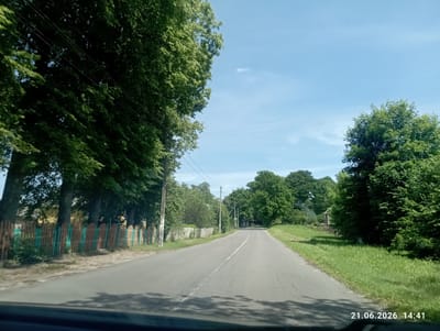 The image shows a paved road leading through a rural area in Хаценчыцкі сельскі Савет, Belarus. The road has a white dashed line down the center. On the left side of the road, there is a wooden fence painted with alternating brown and green sections, behind which are several large trees. On the right side of the road, there is a grassy embankment with more trees. The sky is blue with a few wispy clouds. The date stamp in the bottom right corner reads "21.06.2026 14:41". The view appears to be from a vehicle, indicated by the blurry foreground and the bottom edge of a windshield. There are no people visible in the image.