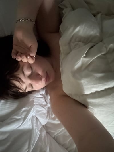 The image displays an individual, appearing female, lying in a bed. The subject has short, dark brown hair and closed eyes, suggesting a state of sleep or rest. The head is turned slightly to the viewer's right. The right arm is raised, with the hand partially covering the forehead. A silver-colored bracelet with heart-shaped links is visible on the right wrist, and a ring is present on a finger of the right hand. The individual's body is partially obscured by white, rumpled bedding, including sheets and a comforter. The lighting in the scene is soft and indirect. No other objects or environmental details are visible beyond the subject and bedding. The image contains no visual information or contextual cues to confirm the specified location of Montepulciano, Italy.
