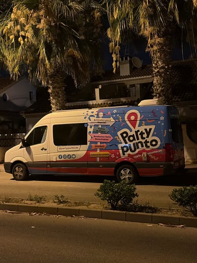 An exterior, eye-level, medium shot captures a white commercial van parked on the side of a street at night. The van is adorned with a vibrant, colorful advertisement for "Party Punto," featuring playful graphics and text. The advertisement spans the entire side and rear of the vehicle, promoting party supplies such as articles for parties, disposable tableware, helium balloons, and confetti. The van has tinted windows and a branding logo that appears to be a red pin icon. Social media icons for Facebook, Instagram, TikTok, and YouTube are visible on the side of the van, along with the website "@partypunto.es". The vehicle is positioned on a paved surface with a curb and a strip of dry grass and sand separating it from a sidewalk. In the background, a residential building with a tiled roof and a satellite dish is partially visible, framed by the fronds of tall palm trees. The scene suggests a commercial operation related to event supplies, likely operating in the city of Orihuela, Spain, given the context. The lighting suggests it is evening or night. No people are visible in the image.