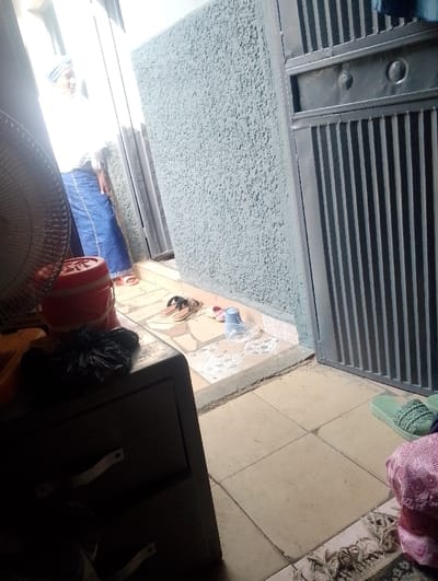 The image captures an interior view looking into a brightly lit doorway or passageway in Dorayi, Nigeria. A person stands within this opening, facing towards the right, wearing a white top, a blue patterned wrapper, and a striped head covering, with light-colored sandals on their feet. The person's expression appears to be a slight smile. To the right of the passageway is a textured light grey wall, beyond which a dark grey metal gate with vertical slats is partially visible. In the foreground on the left, a dark grey cabinet with white handles is partially obscured by a tan fan. A black plastic bag rests on the cabinet, alongside an orange plastic bucket with a white lid. The interior floor consists of light-colored square tiles, with a patterned rug and light green braided slippers visible on the lower right. The ground immediately outside the doorway features different patterned tiles, upon which two pairs of sandals, a light blue plastic cup, and clear plastic material are situated.