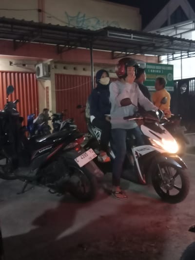 The image captures a scene at night in Sekopek Wetan, Indonesia. Three people are visible, two on a scooter in the foreground and one person in the background to the right. The two individuals in the foreground are seated on a scooter, with the rider wearing a red helmet and the passenger wearing a light-colored face mask and a dark hijab. The rider appears to be gesturing with their right hand. The scooter has its headlight on, illuminating the ground.
In the background, behind the scooter, is a building with red vertical blinds. An air conditioning unit is mounted on the wall. Further in the background, another person wearing an orange shirt is visible. A green sign with white text is mounted on the building, but the text is mostly illegible due to the image quality.
The overall atmosphere suggests a casual, possibly social, gathering or stop. The time of day is clearly night, indicated by the artificial lighting and the dark surroundings. No specific weather conditions are discernible. The presence of scooters and the building's architecture provide some context for a location in Indonesia.