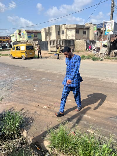 The photo shows a person standing on a street in Ijegun, Nigeria. The person is wearing a blue plaid suit and sandals. The street is paved, with a dirt and grass edge. In the background there are buildings, an auto rickshaw vehicle and other people. There are overhead power lines and a partly cloudy sky.