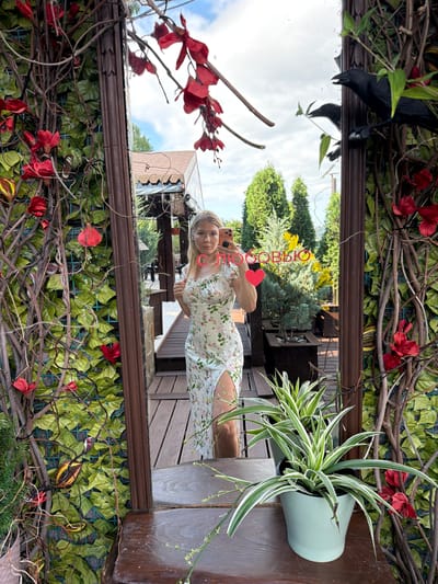 A full-body mirror selfie shows a blonde woman with fair skin and blue eyes standing in front of a decorative wall with artificial greenery and red flowers. She is wearing a light-colored, floral-print dress with a thigh-high slit and a ruffled neckline. She is holding a light orange smartphone in her right hand, positioned to take the photo. The mirror reflects a wooded area with trees, a building with a brown roof, and a cloudy sky. Two black crow figures are perched on the frame of the mirror. In the foreground, a light green pot contains a spider plant. The scene is outdoors, possibly on a patio or balcony, given the wooden decking and potted plants. The woman is in the city of Nizhnyaya Shilovka, Russia.