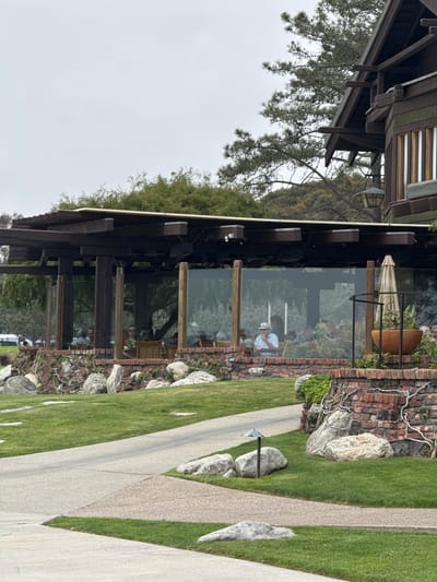 The image depicts an exterior view of a restaurant with a covered patio area in San Diego, California. The restaurant features a dark wood architectural style, consistent with Craftsman or Prairie School design elements, with exposed beams and a sloped roof. The patio is enclosed by glass walls, offering a view of diners seated at tables inside. The patio is supported by wooden posts and the building's foundation appears to be constructed of red brick and large rocks. In the foreground, a manicured lawn with a pathway is visible, interspersed with several large rocks and a small outdoor light fixture. The sky is overcast, suggesting a cloudy day. The overall scene conveys a relaxed and upscale dining atmosphere.