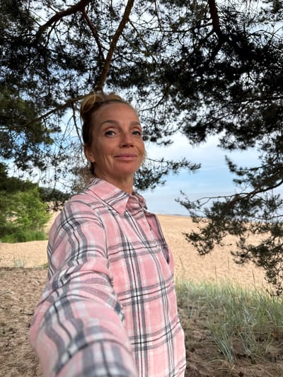 A woman with blonde hair pulled up in a bun, wearing glasses and a pink plaid shirt, stands on a sandy beach under the branches of a pine tree. She is looking towards the camera with a slight smile. The background shows the sea and the sky. The location is Zelenaya Roshcha, Russia.