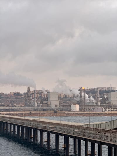 The image depicts a cloudy, overcast day at an industrial complex located in Mers El Hadjadj, Algeria. In the foreground, a long, elevated pier extends over dark blue water. The pier is supported by numerous dark, cylindrical pilings and features a railing with a fence-like structure and a wooden top. Along the pier, pipes are visible, and some lighting fixtures are present.
In the middle ground, the sea meets a shoreline dotted with industrial structures. Several large, white, rectangular buildings dominate this area, alongside a cylindrical white tank. Various pipes and metal frameworks are integrated into the industrial landscape.
In the background, a sprawling industrial facility is situated on a hillside, with numerous tall structures, smokestacks, and buildings. Smoke and steam are visibly rising from several points within the facility, indicating ongoing operations. A prominent feature is a tall structure with a bright orange flame at its apex, suggesting a flare stack. Beyond the industrial area, residential buildings are visible on the upper slopes of the hill, and a power line pylon can be seen further in the distance. The overall atmosphere is industrial and somewhat muted due to the overcast sky.