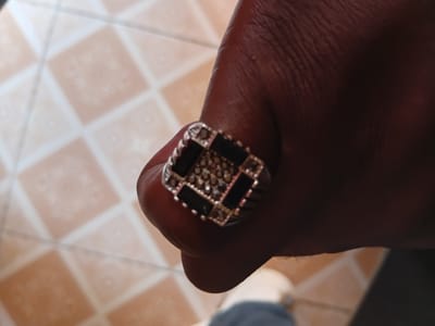 A close-up shot shows a person's hand with a ring on one of the fingers. The ring appears to be made of metal and has a square face with black and silver stones set in a grid pattern. The hand is resting against the person's leg. The background is tiled flooring. The location is unknown.