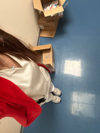 The image depicts an indoor scene, identified as being in the city of Tourcoing, France. A person is visible from the upper torso down to the feet, positioned on the left side of the frame. The individual has long brown hair and is wearing a light-colored, possibly white, collared top. A silver-colored necklace with a light blue pendant is visible. A bright red garment, appearing to be a jacket or sweatshirt, is draped over the person's left arm and shoulder. The person wears white athletic shoes with light-colored soles. They are standing on a clean, medium-blue floor, which shows reflections of overhead lighting and minor scuff marks. Against a light-colored wall to the left, three brown cardboard boxes are present. One open box is situated near the person's feet. Further along the wall, two additional boxes are stacked; the top box is open and contains multiple small, individually packaged items, while the bottom box is closed.
