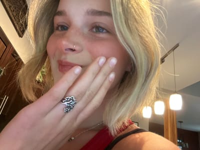 A close-up, inverted selfie captures a person with blonde hair, fair skin, and blue-grey eyes, appearing to look upwards towards the viewer. The image is rotated, presenting the subject upside down. Their right hand is raised, with fingers gently resting across their upper cheek and partially obscuring their mouth and nose. A small silver hoop nose ring is visible in their left nostril.
The person is wearing a red top or bra strap, visible at the edge of the frame, and a thin silver chain necklace with a small bead is around their neck. A distinctive silver ring, featuring an intricate, organic, or tree-like design, adorns their right ring finger. Their blonde hair, highlighted by light, frames their face.
In the background, out of focus, are three bright, glowing white pendant lights or lamps, indicating an indoor setting. A wooden structural beam or post is visible behind these lights. Further in the background, glimpses of dark wooden cabinetry and a framed decorative piece with an intricate pattern can be seen. Another simple wooden hanging decoration is visible on a wall. The overall lighting suggests an interior space, possibly in the evening or an area reliant on artificial illumination. The activity is a posed selfie, capturing a direct gaze despite the unusual angle and partially obscured face. This image is set in Whistler, Canada.