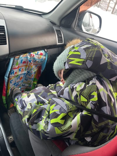 Within the front passenger seat of a vehicle, a young individual is observed in a curled, resting position. The exterior environment, visible through the windows and side mirror, is a snow-covered landscape under an overcast sky, with the location attributed to Италмас, Russia. The individual is dressed in a heavily insulated winter suit, predominantly black, grey, white, and neon yellow-green, featuring geometric patterns and the word "BROOKLYN". A light blue knitted beanie with a fur pom-pom covers the head, complemented by a matching knitted neck gaiter. Black boots are visible on their feet. The individual's feet are braced against the lower section of the dark grey dashboard, partially obscuring a colorful fabric organizer adorned with Cyrillic alphabet characters and illustrative images. A black seatbelt strap crosses the individual's chest. The vehicle interior shows dashboard air vents.