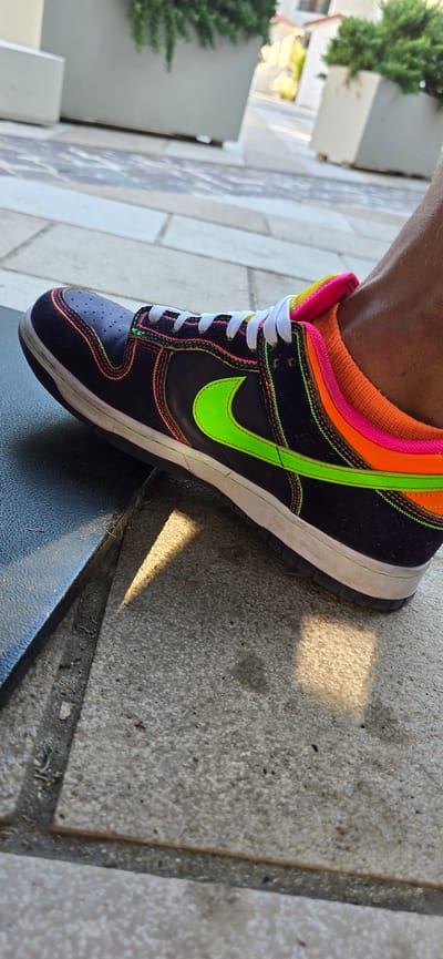 A close-up shot captures a single Nike sneaker, a dark purple low-top with vibrant neon green accents, including the signature Nike swoosh. White laces are threaded through the eyelets, and the trim of the shoe features neon pink and orange stitching. The sneaker is worn by a person whose skin is visible around the ankle, with an orange sock peeking out. The shoe rests on a concrete surface outdoors, with dappled sunlight creating shadows. In the background, blurred greenery and light-colored planters suggest a park or public space, consistent with an outdoor setting in Radovići, Montenegro.