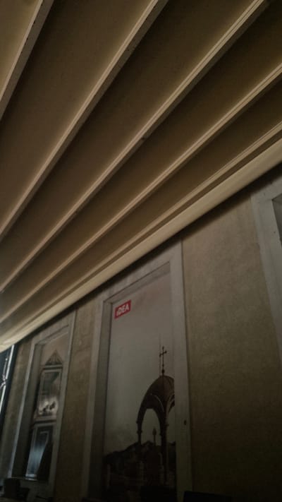 The image depicts an interior view of a building, likely an institutional or public space, with a distinctive ceiling structure. The ceiling consists of parallel, angled beams, casting shadows and creating a ribbed effect. The walls are textured in a neutral, earthy tone.
On the wall, there are two framed areas. The one on the left appears to be a window or a decorative panel with a dark frame and what looks like a reflection or a dark image within. The larger framed area to the right is a mural or a large poster. This mural features an architectural element, possibly a dome or a cupola with a cross at its apex, set against a dark, possibly hilly landscape. A red rectangular sticker with the word "IDEA" in white text is affixed to the upper portion of this mural.
The overall lighting is dim, with the angled ceiling beams being the brightest elements, suggesting an artificial light source positioned above and to the side. The scene appears to be unoccupied by people. The architectural style and the mural suggest a historical or cultural context.
While the image itself does not contain specific geographical markers, the prompt specifies the location as Cetinje, Montenegro. Therefore, this scene is located within Cetinje, Montenegro. The specific building or room is not identifiable from the visual information alone.