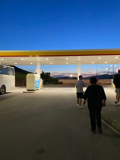 The image depicts a gas station, identifiable by the Shell logo and gas pumps, likely in Kalotina, Bulgaria. The sky transitions from blue to hues of sunset near the horizon. Three individuals are present. One wears a light-colored shirt and dark shorts, while another wears a black jacket and dark pants. The third individual is partially visible in the top-left corner. A white bus is parked near the gas pumps.