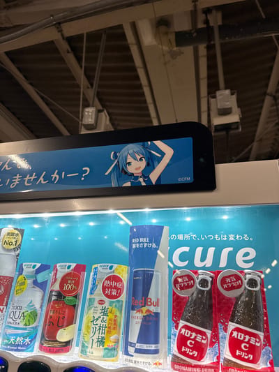 This media file shows the interior of a brightly lit vending machine, likely in a public space given the presence of product packaging. The primary focus is on the arrangement of various drinks. Two bottles of "Oronamin C Drink" are prominently displayed near the top, each with "元気ハツラツ!" (Genki Hatsu Ratsu! - Energetic and Lively!) written on its cap. Below these, a can of "Red Bull" with the slogan "翼をさずける。" (Tsubasa wo Sazukeru - Gives You Wings) is visible. Further down, there are several "Acure made" branded drinks. One features "Salty Citrus Jelly" with "河内晩柑" (Kawachi Bankan - a type of citrus fruit) and "伯方の塩" (Hakato no Shio - Hakata Salt). Another displays "ストレート果汁100%" (Straight 100% Fruit Juice) with an image of an apple and "青森りんご" (Aomori Ringo - Aomori apple). The bottom visible drink is "天然水" (Tennen Sui - Natural Water) branded "AQUA". A prominent blue panel on the left side of the vending machine features an anime-style illustration of a girl with blue hair and headphones, along with Japanese text that appears to be a question. The overall impression is one of a well-stocked vending machine offering a variety of beverages, likely in a location where such a selection would be appreciated, such as a train station or a convenience store, given the context of Matsudo, Japan. The lighting suggests it could be daytime or a well-lit indoor environment.