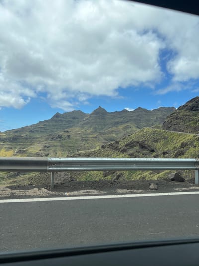 A daytime, rotated view shows a landscape taken from a moving vehicle, specifically in Mogán, Spain. The sky features blue patches with white clouds. Adjacent to the sky, rugged, green-covered mountains rise sharply. A roadside barrier runs vertically through the image, separating the mountainous landscape from the road surface, which takes up the right side of the image. The view is partially framed by the vehicle's interior structure.