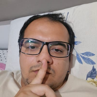 A man wearing glasses and a light-colored t-shirt is lying on a pillow with a floral pattern. His right index finger is held to his lips, signaling silence. The man's eyes are open and looking towards the camera. The image appears to be a selfie, taken from a close-up perspective. The background is a pillow with blue and white floral designs. The setting is indoors, possibly a bedroom. There is no information in the image that indicates the location is in Küçükçekmece, Turkey.