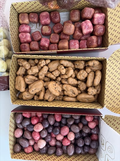 The image shows three open boxes of assorted candies. The boxes are stacked vertically. The top box contains irregularly shaped candies with a pinkish-red hue. The middle box contains candies coated in a light brown, caramel-like substance, with some appearing to be nuts or dried fruits. The bottom box is filled with round candies in various shades of pink, purple, and dark brown, resembling chocolate-covered berries or nuts. The boxes are decorated with a geometric pattern. The overall setting appears to be indoors, possibly a shop or market stall, and the candies are arranged for display. The location is stated to be the city of Holon, Israel.