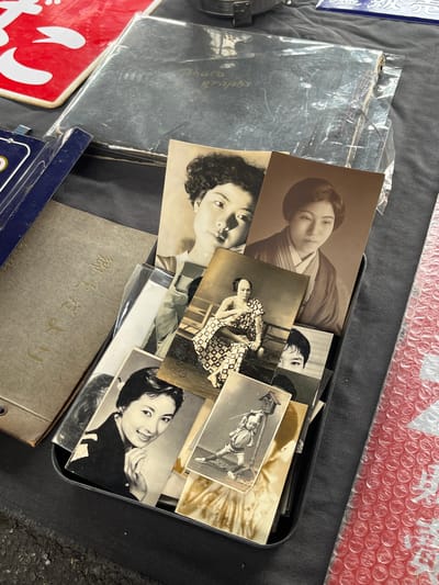 A high-angle shot depicts a collection of black and white photographs inside a black metal tray, interspersed with a small gray book with Japanese text. The photographs appear to be portraits of Japanese women, and the composition is cluttered. Other items, including red and blue signs, clear plastic coverings, and bubble wrap, are adjacent to the tray. The surface the tray sits on is a dark gray color. The photographs are old, and the gray book appears to have a metal rivet on its binding. The location may be Shinagawa, Japan, suggested by the Japanese text on the book.