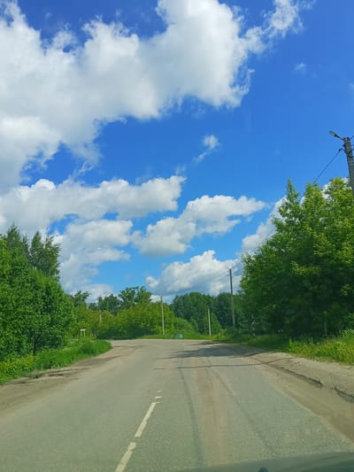 The image displays a bright, sunny view of a rural or semi-rural asphalt road, presumably in Teykovo, Russia, as provided by the context. The road curves gently to the right in the mid-ground, leading the eye into the distance. A faded white dashed line is visible on the road surface in the foreground, indicating a two-way traffic path.

Dense, lush green deciduous trees and bushes line both sides of the road, suggesting a summer season. The foliage is thick and vibrant, forming a natural corridor. The road's shoulders are unpaved, consisting of dirt, gravel, and sporadic patches of grass and weeds.

Above the tree line, a vast expanse of brilliant blue sky is filled with numerous fluffy, white cumulus clouds, indicating fair weather conditions. Several utility poles, carrying power lines, run parallel to the road on both sides, stretching into the background. One prominent pole on the right features a modern, dark-colored streetlight fixture. On the left side of the road, behind some bushes in the mid-distance, a small, white rectangular sign is visible, but any text on it is illegible due to resolution and distance.

The bright, even lighting and clear sky suggest it is daytime, likely mid-morning or early afternoon. The scene is devoid of any visible people or vehicles other than the implied presence of the photographer's vehicle, conveying a sense of peaceful travel through a verdant landscape.