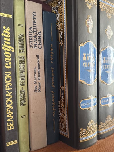A bookshelf displays a row of books, primarily in Cyrillic script, on a wooden shelf. The books are of varying heights and cover colors, including black, light green, beige, and dark blue. The spines of the books are facing the viewer. The leftmost black book has yellow lettering that appears to read "БЕЛАРУСКА-РУСКИ СПОУНIK". The second book, light green, has yellow lettering that says "РУСКО-БЕЛОРУССКИЙ СЛОВАРЬ". The third, beige book, has black lettering that includes "УЛИЦА МЛАДШЕГО СЫНА". The dark blue book next to it has yellow lettering, possibly in Cyrillic. The remaining books are black with ornate gold and blue designs on their spines. The text on these spines appears to be in Cyrillic, with blue labels that read "ИЗБРАННЬІЕ ЖИТИЯ СВЯТЬІХ" and also some months like "июль-". The image is taken from a slightly high angle, looking down at the books. The lighting suggests an indoor environment, likely a home or library. The background is out of focus, but appears to be a continuation of the bookshelf. There are no people or animals present in the image. The scene is static and orderly. The specific location in Chachersk, Belarus, cannot be determined from the visual information alone, as the image depicts only a bookshelf and its contents.