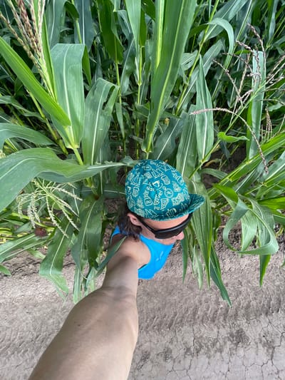 A person wearing a teal hat with various white icons and a blue tank top stands in a cornfield in Perevoznoe, Russia. The person is wearing sunglasses and their arm is extended, holding the camera. The corn stalks are tall and green, with some tassels visible. The ground is dry and cracked.