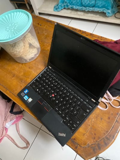A ThinkPad laptop is open on a wooden table. A clear plastic container filled with food sits to the left of the laptop. Several items, including what appears to be a bag, are visible around the table. The table itself shows signs of wear, and the background includes a tiled floor and various objects. The laptop screen is black.