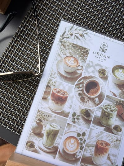 This media file shows a close-up of a menu for "URBAN GARDEN" cafe, possibly located in La Vila Joiosa, Spain. The menu features a collage of high-quality photographs showcasing various coffee and matcha drinks, artfully presented with latte art and garnishes. The aesthetic is clean and minimalist, with a focus on natural elements like olive branches and dried flowers, suggesting a tranquil and sophisticated atmosphere.
The menu is laid out on a dark woven placemat, with a pair of modern sunglasses with gold frames resting on top. The lighting appears to be natural, hinting at daytime. The overall impression is one of a relaxed, stylish cafe experience, inviting customers to enjoy a moment of peace with a beverage. The menu itself has "URBAN GARDEN" written in large, capitalized letters, with a stylized leaf logo above it. The background of the menu is a subtle, textured paper. The image conveys a sense of calm and indulgence, typical of a cafe setting.