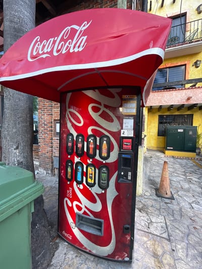 The image shows a weathered Coca-Cola vending machine in Playa del Carmen, Mexico. The machine is red and features the Coca-Cola logo prominently, with prices for beverages ranging from $20 to $40. A red Coca-Cola branded awning extends over the machine. The machine is positioned on a stone-paved area, next to a green waste container. Behind the machine, there is a yellow building, a brick wall and a cone.