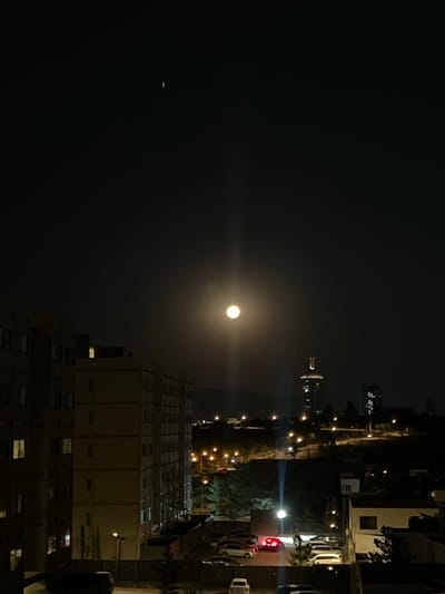 The image depicts a nighttime urban landscape in León, Guanajuato, Mexico, captured from an elevated perspective. The sky is dark, featuring a bright, full moon positioned centrally, emitting a distinct vertical light ray. A faint, crescent-shaped object is visible in the upper left quadrant of the sky.
In the left foreground, a multi-story, light-colored residential building occupies a significant portion of the frame, with several windows displaying interior illumination. Below this building, a ground-level parking area is visible, containing multiple parked vehicles; one car's red taillights are illuminated.
The midground reveals a city sprawl characterized by numerous streetlights and illuminated buildings. A prominent, tall building with a distinctive illuminated crown, identified as the Poliforum León Tower, stands slightly to the right of the image center. Other smaller buildings and patches of vegetation are also visible. Distant hills or mountains are faintly discernible on the horizon line. No people are visible.