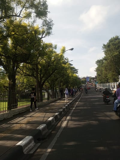 The image depicts a street scene with pedestrians and vehicles. A line of people walks along the sidewalk on the left side of the road, protected by a low fence. The sidewalk is shaded by trees. On the right side of the road, people are riding motorcycles. The road has a white dividing line. Streetlights are visible along the road. In the background, there are buildings, a large sign, and a bridge or overpass. The sky is partially cloudy. The location is unknown but the scene is not in Phou Khoun, Laos.