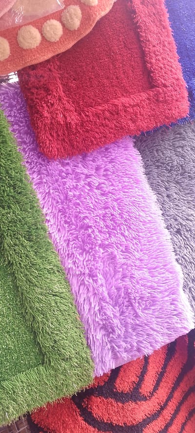 The scene, located in Okere-Urhobo, Nigeria, displays a close-up, overhead view of multiple overlapping textile floor coverings. The primary subjects are several shaggy rugs or mats of varied colors and textures. From top to bottom, partially visible objects include an orange mat with five light-colored, spherical pom-pom-like embellishments along its edge, layered over a rectangular deep red shaggy mat. Below this, a bright lavender-purple shaggy mat is visible. To the left, a vibrant green mat with a short, dense pile resembling artificial turf is positioned diagonally. To the right, a segment of a grey shaggy mat is discernible. At the bottom, a red mat featuring prominent black, wavy, stripe-like patterns is partially visible. The objects are static, without any visible people or interactions. The immediate environment is obscured by the close focus on the textiles, suggesting a display setting.