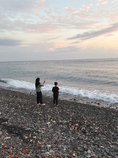 Two people stand on a pebble beach in Abang, Indonesia, facing the ocean. The person on the left, wearing a hijab and a green shirt, holds up a phone to take a picture. The person on the right, wearing a black t-shirt, stands with their back to the camera. Waves crash gently on the shore, and the sky is a mix of soft blues and pinks, suggesting either sunrise or sunset. The beach is covered in dark gray pebbles and some debris.