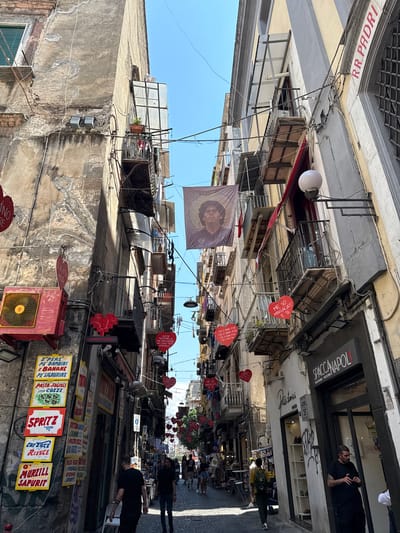 This is a street view in Naples, Italy, with the image oriented vertically. The scene depicts a narrow alleyway lined with buildings on both sides, characteristic of the Spaccanapoli area. The sun is shining, indicating daytime.
Several people are visible, some walking down the street, others appearing to be shopkeepers or patrons. The buildings are old and have a weathered appearance, with balconies and windows. Many wires and electrical conduits are strung across the alley, typical of older European cities.
Various objects are displayed, including signs advertising food and drinks such as "SPRITZ," "PASTA FAGIOLI CON COZZE," and "PASTA PARTE PROVOLA SPIZIOSA." Some signs are heart-shaped and have Italian text. A large banner with a portrait, possibly of a local personality, hangs in the middle of the street. Another sign reads "SPACCANAPOLI" and "CUOREDINAPOLI," confirming the location. A red fan unit is also visible.
The overall impression is a lively, bustling street scene in a historic Italian city, filled with local character and commerce. The perspective looking up the alley emphasizes the tall buildings and the sky above.