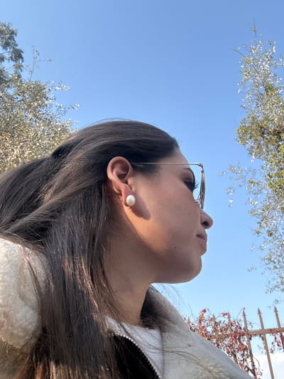 A woman's low-angle portrait shows her looking up at a clear blue sky partially obscured by tree branches and a structure. She wears glasses pushed slightly on her face and a white pearl earring. Her brown hair cascades around her face. She is wearing a white, textured jacket with a visible zipper. A shadow falls across her face.