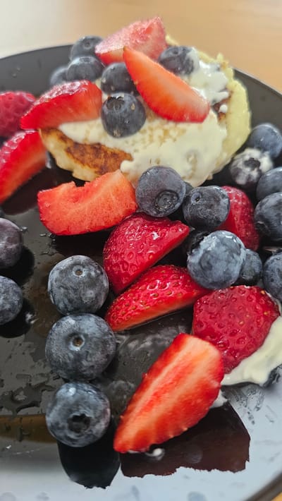 A close-up, overhead shot shows a dark plate filled with pancakes topped with cream and surrounded by fresh blueberries and sliced strawberries. The plate is wet, with water droplets visible, suggesting it may have been rinsed or is being served with a syrup. The background is blurred and appears to be a wooden surface. The scene is an indoor setting, likely a kitchen or dining area. There are no people or animals visible in the image. The location is not specified as Slengiai, Lithuania, as there is no information to support this.