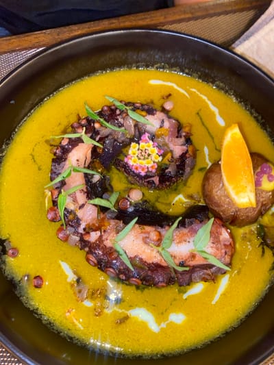 A close-up, top-down view shows a prepared dish served in a black, round ceramic bowl, likely in a restaurant setting in Dikwella, Sri Lanka. The dish features cooked octopus tentacles arranged in a U-shape within a vibrant yellow-orange, creamy sauce or purée. The octopus pieces are dark reddish-brown with lighter pinkish-white sections, exhibiting visible suckers and some charred edges. They are garnished with small, pale green leaves and, at the top center, a cluster of tiny multi-colored flowers in shades of yellow, pink, and white. To the right of the octopus, a small, whole roasted potato is present, topped with a bright orange slice and a purple garnish. The yellow-orange sauce has visible specks and a few white, streaky drizzles. In the blurred background, a warm-toned wooden surface and a textured fabric item are discernible, along with a dark, indistinct shape that may be a person's clothing or arm.