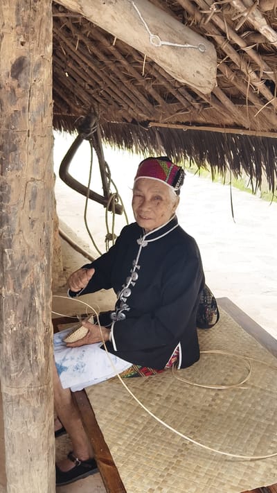 An elderly Asian woman is depicted weaving under a thatched shelter in Jiyang District, China. She sits on a light-brown, woven mat with a square pattern. The woman wears a black, long-sleeved jacket featuring white trim and a vertical line of approximately eleven large, circular, silver-toned fasteners. Her head is adorned with a colorful, patterned headpiece, primarily red, pink, and green, displaying geometric designs. A white cloth is draped over her lap. She holds a small, light-colored, partially woven basket or container, actively manipulating thin strips of weaving material. Additional strips are coiled on the mat beside her. Her gaze is directed towards the viewer, exhibiting a slight smile. To her left, a substantial, weathered wooden pillar supports the shelter's roof, which is constructed from dried leaves or straw layered over wooden beams. In the background, behind the woman, a dark, rustic wooden structure with attached ropes is visible, beyond which lies a light-colored ground area. She wears black flat shoes with a single strap.