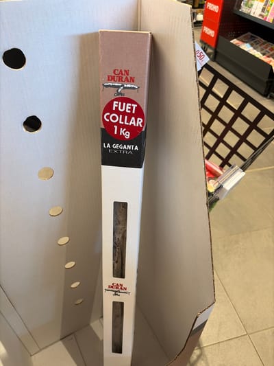 The image shows a package of "Can Duran Fuet Collar 1 Kg La Geganta Extra" sausage product in a cardboard box. The box has several circular ventilation holes. A price tag for 0.50 Euros is visible, as is a "PROMO" sticker. The background suggests a store environment, likely a supermarket or grocery store. The city of Dinant, Belgium, is not discernible in the image.
