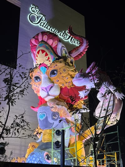 The image displays a large, vividly illuminated, multi-tiered sculpture erected outdoors at night, positioned against the facade of El Palacio de Hierro department store in León, Mexico. The sculpture itself is a stack of fantastical creatures, reminiscent of *alebrijes*, featuring a prominent red and pink horned figure at its apex, with an open mouth displaying jagged white teeth. Directly below this is a jaguar-like head with orange spotted fur, large blue eyes, and a pink nose. Further down, a light grey figure with a blue heart-shaped jewel on its forehead is visible, followed by a scaled blue creature at the sculpture's base. The modern, light-colored building in the background prominently features the illuminated cursive sign "El Palacio de Hierro." Bare tree branches partially obscure the view in the foreground. Yellow metal scaffolding and a ladder are visible on the right side of the sculpture, suggesting ongoing installation or maintenance. The scene is brightly lit, contrasting with the dark night sky.