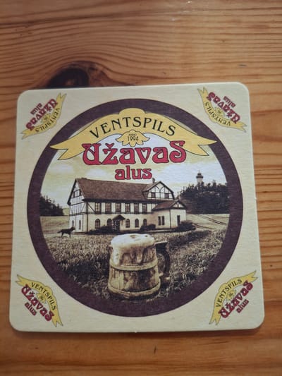 The image is a close-up, overhead shot of a round beer coaster placed on a wooden surface. The coaster features a sepia-toned illustration depicting a building that resembles a historical brewery or inn with a timber-framed upper story. In front of the building, a field is shown with a dog in the middle ground and a frothy beer mug in the foreground. The background includes trees and a distant lighthouse. The coaster has a decorative border with text. The text on the coaster, which appears to be in Latvian, reads "VENTSPILS DZAVAS ALUS" and "ANNO 1994". The coaster is positioned on a wooden table with visible grain patterns.