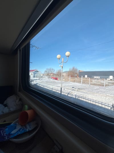 The image captures an exterior view through a window, likely from a train, in the city of Chany, Russia. The foreground shows a dark, textured interior surface with various objects: an orange cylindrical cup lying on its side, a white bowl, a blue and white wrapper (partially visible, displaying "ОКО"), a green cap, and crumpled white paper.
Beyond the window, a sunny, clear blue sky dominates the upper portion of the frame. The ground is characterized by patches of snow and bare dirt, indicating a cold season. Train tracks are visible, partially covered by snow, running parallel to a light-colored metal fence that extends across the mid-ground. A prominent silver lamppost with two spherical, opaque white light fixtures and a small dark camera attached to its pole stands left of center.
In the background, on the left, a small light-grey building with a red pitched roof is visible. To the right, a large, dark grey industrial-style building with a flat roof and several white rooftop ventilation units is present. Further back, a multi-story building with multiple windows can be seen, partially obscured by several bare deciduous trees. Thin dark power lines traverse the clear sky. No individuals are visible in the scene. The setting appears to be a train station or railway area in Chany, Russia.