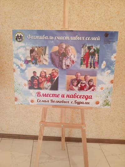 The image shows a poster displayed on a wooden easel. The poster features multiple photographs of families and text in Russian. The title of the poster, "Фестиваль счастливых семей" (Festival of Happy Families), is prominently displayed. Below this, there is another line of text, "Вместе и навсегда" (Together Forever), followed by "Семья Волковых с.Буралы" (Volkov Family, village Buraly). The photographs depict various family groups, including adults and children, in indoor and outdoor settings. The poster also includes a logo and information about the Presidential Grants Foundation. The background of the poster is a light blue sky with images of daisies. The easel is placed on a tiled floor in front of a textured wall. The overall scene suggests an event or celebration related to families.