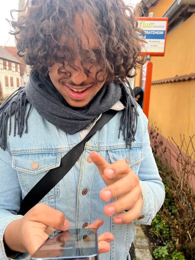 A male individual is depicted in a close-up, outdoor scene at a bus stop in Meistratzheim, France. The subject, positioned centrally, has dark, curly hair obscuring most of his forehead and eyes. He is smiling, with a mustache and his mouth slightly agape, revealing teeth. He wears a light blue denim jacket with copper-colored buttons and a dark grey fringed scarf. A black strap, likely from a bag, crosses his chest diagonally. He holds a dark smartphone horizontally in his left hand, its screen reflecting ambient light, while his right hand is poised above the device with fingers splayed.
In the upper right background, a blue and white "Fluo GrandEst" bus stop sign is clearly visible, displaying "MEISTRATZHEIM" at the top, with "Strasbourg" and "Erstein" listed below. Behind this sign, an ochre-yellow building with a tiled roof is present. To the left of the main subject, a traditional half-timbered building with light-colored walls and dark wooden beams is partially visible. Sparse, dry vegetation is visible in the lower right and mid-right background. A second, out-of-focus individual, clad in dark clothing, is partially visible behind the main subject's right shoulder near the bus stop sign. The scene is illuminated by diffused daylight, indicating an overcast sky.