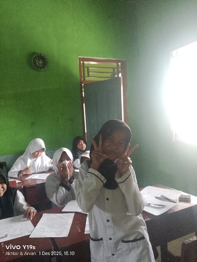A daylight image, captured on December 1, 2025, at 15:10, depicts an interior classroom scene within the city of Wanguk, Indonesia. The room features textured green walls and multiple individual wooden desks. On the right side of the frame, a window with a brown wooden frame is heavily illuminated by an external light source, causing significant overexposure. A partially open dark-colored door, also with a brown wooden frame, is visible adjacent to the window. On the upper left wall, a circular, dark-colored decorative emblem is mounted.
Five female individuals, identified as students, are present within the frame, most seated at desks. The prominent subject, positioned right of center in the foreground, wears a black hijab and a white long-sleeved uniform with black pocket and cuff trim. She faces the camera, smiling, with both hands raised, forming "peace" signs. Her face is brightly lit, resulting in some overexposure. To her immediate left, a female student wearing a white hijab sits at a desk, holding a white tissue to her nose. Further left, another female student in a white hijab is seated, looking down at papers placed on her wooden desk. In the background, a fourth female student wearing a black hijab is visible, seated at a desk. Papers and writing instruments are distributed on the wooden desk surfaces.