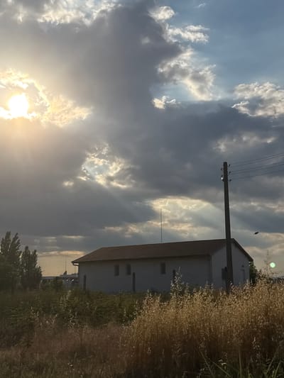 The image depicts a dramatic sky with sunlight breaking through dark, stormy clouds. The sun, a bright orb, casts rays that illuminate parts of the clouds, creating a contrast between light and shadow. In the foreground, to the right, is a building with a reddish-brown roof and a light-colored facade. It appears to be a house or a small commercial building. Tall, dry grass or reeds are visible in front of the building, adding a natural element to the scene. A utility pole with power lines extends across the frame, connecting to the building. The overall impression is one of natural power and the changing atmosphere, possibly indicating the end of a storm or an approaching one. The time of day is likely late afternoon or early evening, given the angle of the sun and the warm tones in the sky. The location is Gelemenovo, Bulgaria, which is not explicitly indicated by any text or visual cues in the image itself. There is no visible text in the image. The scene evokes a sense of awe and perhaps a touch of melancholy due to the moody sky.