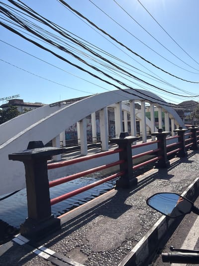 The image shows a concrete bridge with a white arch structure over a body of water. The bridge has a railing with black posts and red bars. The water reflects the sky and the bridge structure. On the left side of the image, a building with a sign that reads "SINGARAJA HOTEL" is visible. Overhead, a tangle of electrical wires crisscrosses the bright blue sky. The foreground shows a textured pavement surface and a motorcycle mirror reflecting the scene. The context of Singaraja Hotel places this scene in the city of Buleleng, Indonesia.