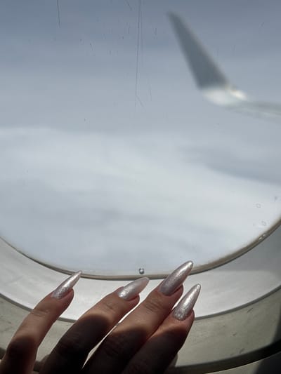 The image shows a close-up of a person's hand with long, pointed, light-colored, glittery nails resting on the edge of an airplane window. The window frame is white and rounded, and the view outside appears to be clouds, with a blurred airplane wing visible in the upper right portion of the frame. The lighting suggests it is daytime. The scene is consistent with being inside an airplane. No specific location within the city of Antung Vien, Cambodia can be determined from this image.