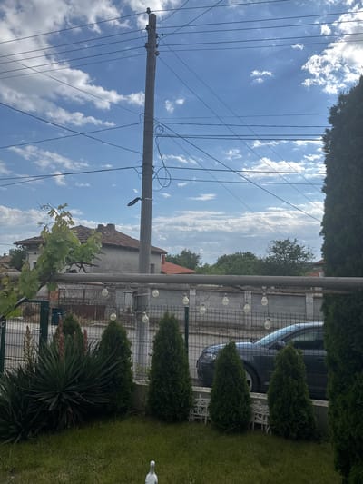 The image depicts an outdoor scene in Kardam, Bulgaria. A concrete utility pole with numerous electrical wires dominates the left side of the frame. The sky is blue with scattered white clouds. To the right of the pole, a residential area is visible, including a house with a red-tiled roof and a gray wall. A metal fence runs parallel to the house, decorated with string lights. Behind the fence, a dark-colored car is partially visible. A row of cone-shaped evergreen trees and a spiky yucca plant line the edge of a grassy area on the right side of the image. A white bird is on the grass in the right corner. Vine foliage is visible in the foreground.